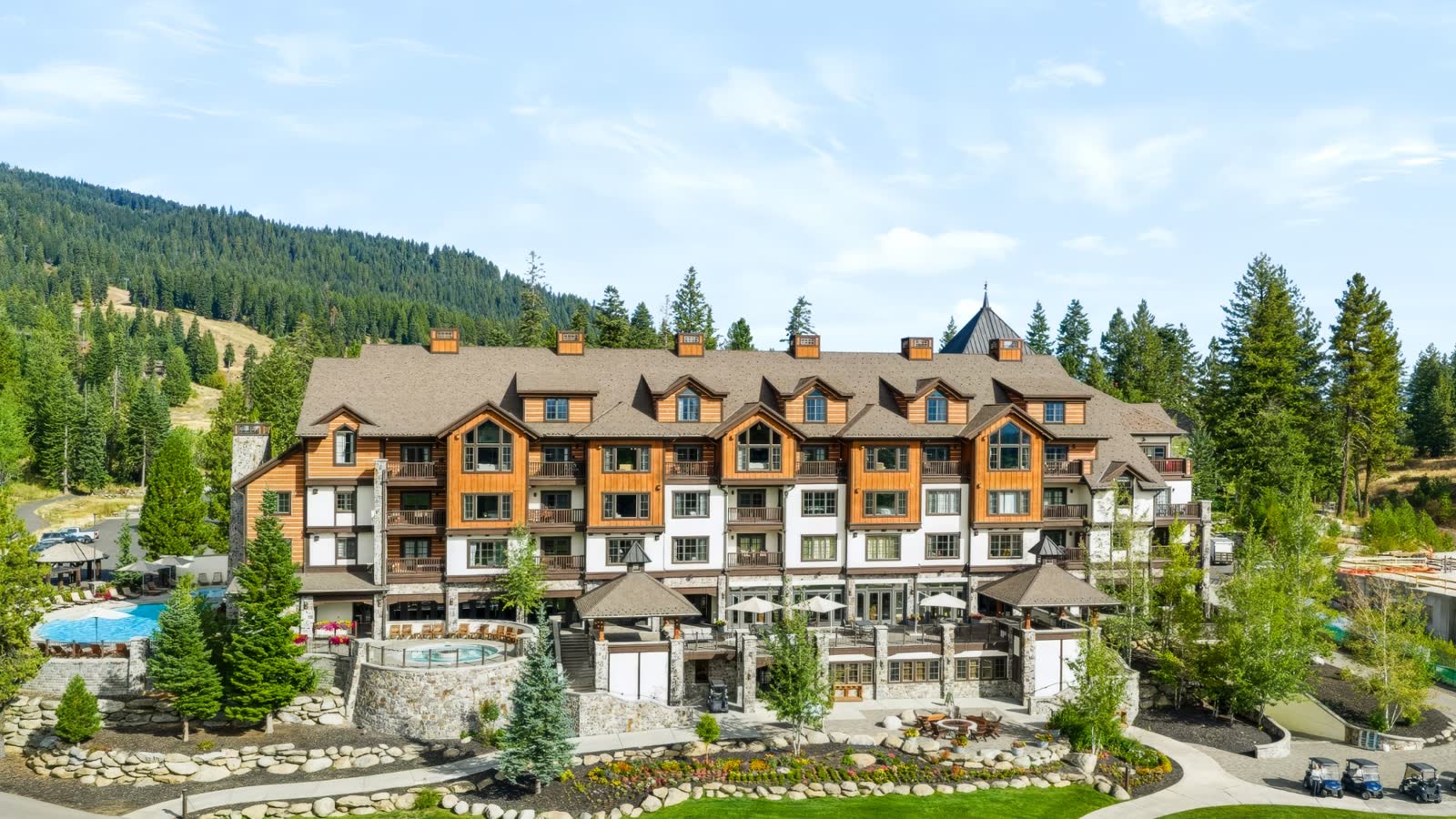 Aerial view of an alpine lodge with pool, hot tub, and landscaped grounds beneath a forested mountain