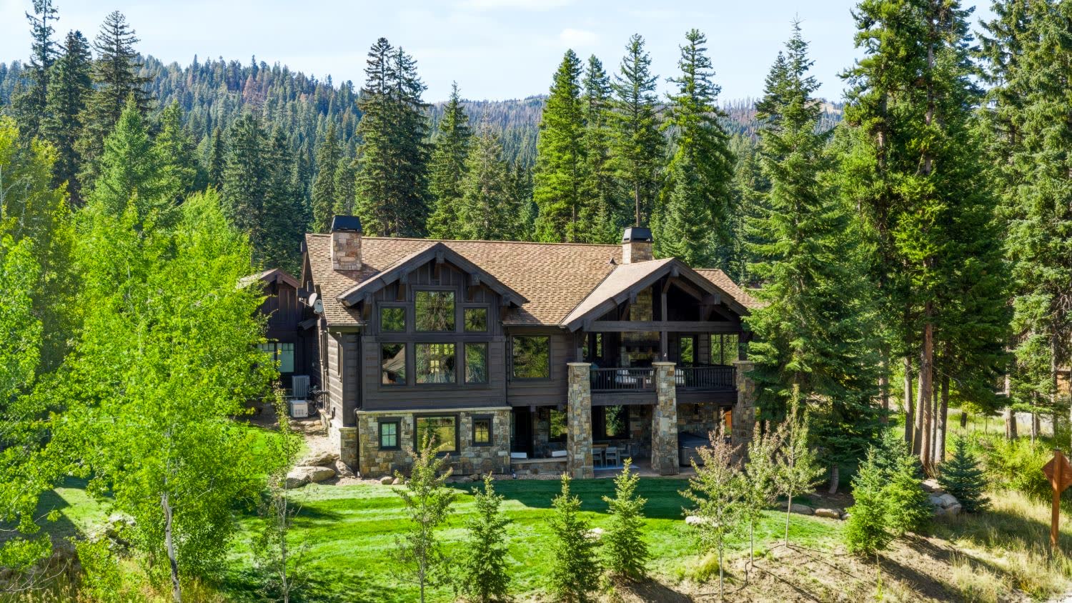 Aerial view of a timber and stone mountain home on a green lawn surrounded by evergreen forest